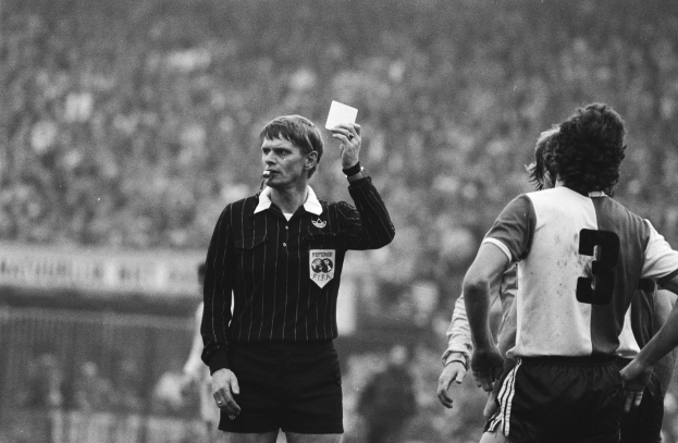 Ein Schiedsrichter in einem Schwarz-Weiß-Bild hält eine rote Karte vor einer Menge von Menschen in Fußballtrikots, mit einem Zaun im Hintergrund.