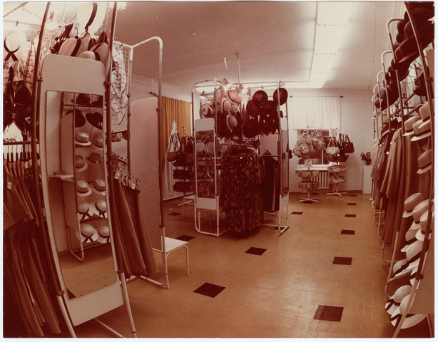 Ein Schwarz-Weiß-Foto eines alten Kleiderladens mit Kleiderstangen, Hutständern, einem Spiegel, einem Stuhl, einem Tisch mit Gegenständen, Vorhängen und Deckenleuchten.