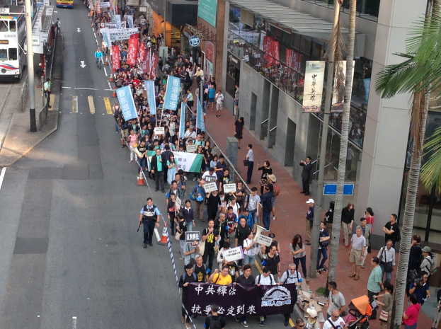 Eine große Gruppe von Menschen marschiert auf einer Straße in Hong Kong, hält Schilder und Plakate hoch, mit Bäumen und Glasfassadengebäuden, Fahrzeugen und Hinweistafeln auf beiden Seiten.