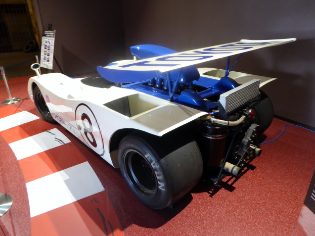 Ein weiß-blauer Rennwagen in einem Museum ausgestellt, umgeben von Absperrungen und Informationsschildern, mit einer Wand im Hintergrund, die auf das Goodwood Festival of Speed hindeutet.