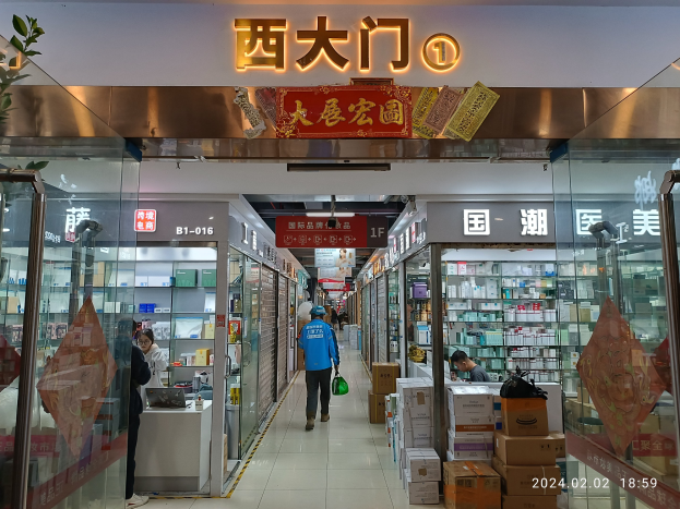 Innenansicht einer chinesischen Apotheke mit Glaswänden, Regalen voller Artikel, verstreuten Kartons, einer Pflanze links und einem Namensschild oben.