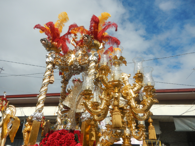 Ein prunkvoller gold- und rotgeschmückter Festwagen mit Blumen und anderen Dekorationen bei einem Karnevalsumzug, im Hintergrund ein Gebäude, Strommasten mit Drähten und ein bewölkter Himmel.