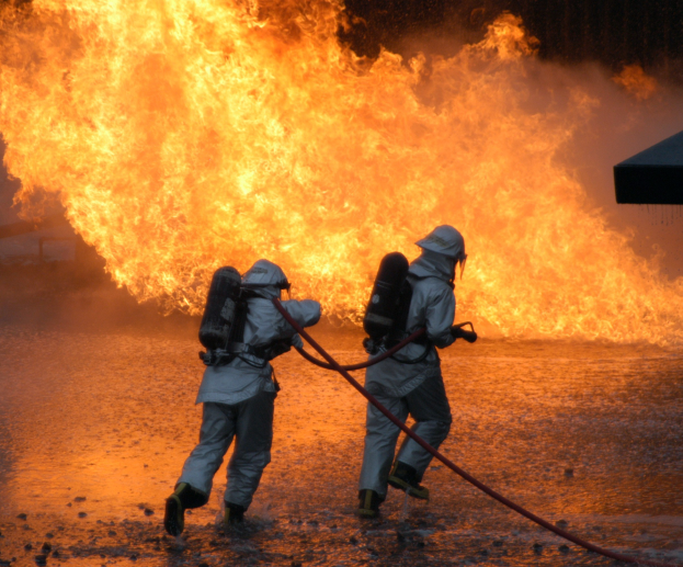 Zwei Feuerwehrleute in Schutzausrüstung sprühen Wasser aus einem Schlauch auf ein Feuer im Hintergrund, mit einem Objekt auf der rechten Seite.