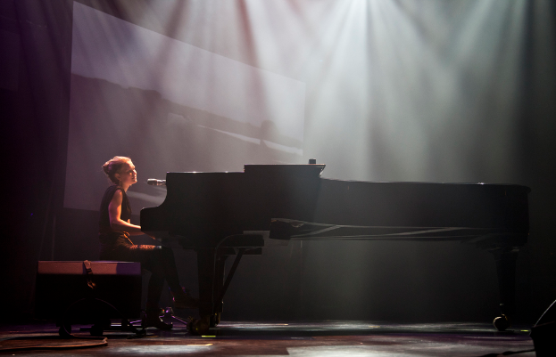 Eine Frau in einem Kleid sitzt an einem Piano mit einem Mikrofon davor, unter einem zentralen Licht, mit Rädern darunter.
