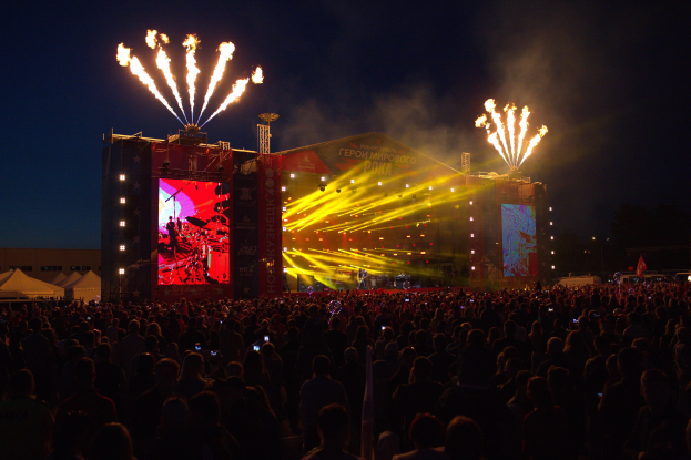 Menschenmenge auf einem Musikfestival mit beleuchteter Bühne und Feuerwerk am dunklen Himmel.