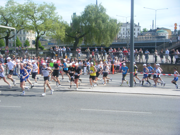 Eine Gruppe von Menschen, die bei einem Marathon auf einer Straße mit einem Tor, einer Ziellinie, Absperrungen, Pfosten, Schildern, einer Brücke, Gebäuden, Bäumen und einem bewölkten Himmel laufen.