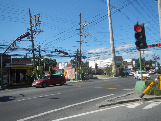 Ein rotes Auto fährt an einer Ampel vorbei auf einer Straße, die von Strommasten und Drähten gesäumt ist, mit Gebäuden, Bäumen und einem klaren blauen Himmel im Hintergrund.