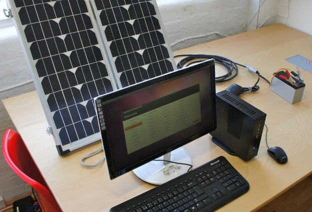 Ein Computersystem mit Tastatur, Maus, CPU, Solarpanel, Batterie, Chips und Kabel auf einem Tisch, mit einem roten Stuhl und einer weißen Wand im Hintergrund.