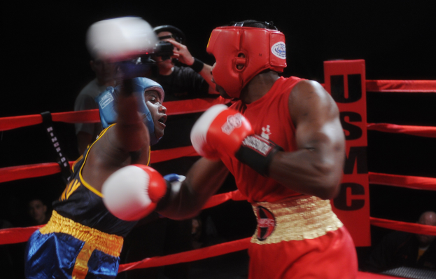 Zwei Boxer im Ring bei einem Kampf, mit einem Fotografen und Zuschauern im Hintergrund bei gedämpfter Beleuchtung.