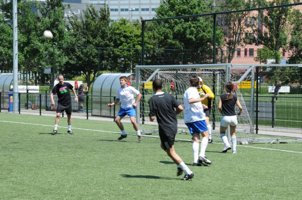 Personen in Kleidung beim Fussballspielen auf einem Feld mit einem Zaun, einem Tor und einem Pfosten mit einem Fussball oben links, mit Bäumen und Gebäuden im Hintergrund.