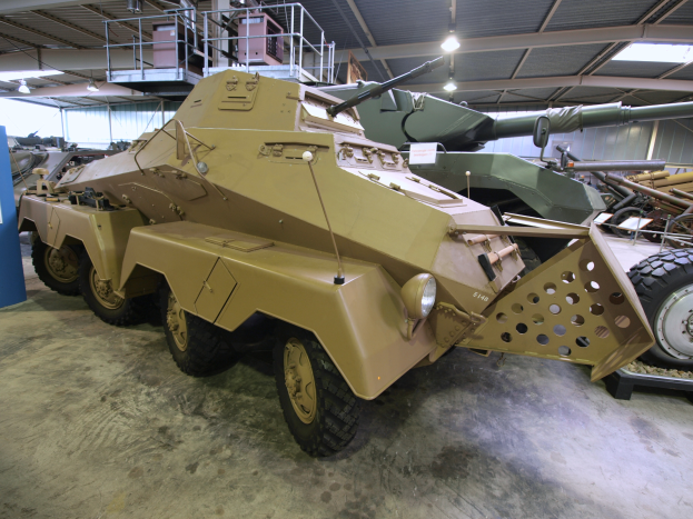 Ein gepanzertes Mannschaftsfahrzeug steht in einem Gebäude, umgeben von anderen Militärfahrzeugen, mit einer Tafel mit Text links und Geländern und Lampen im Hintergrund.