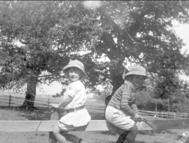 Zwei junge Jungs in Hüten mit Baseballschlägern in einem grünen Park mit Bäumen und einem Zaun im Hintergrund, schwarz-weiß.