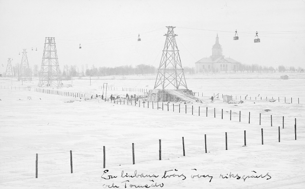 Schwarze-Weiß-Foto eines Skilifts in einem verschneiten Feld mit Stützpfählen, Oberleitungen, Bäumen und einem Gebäude im Hintergrund; Text unten.