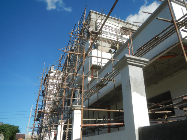 Ein unter Baugerüst stehendes Gebäude, umgeben von Bäumen, Pfählen und Drähten, unter einem bewölkten Himmel.