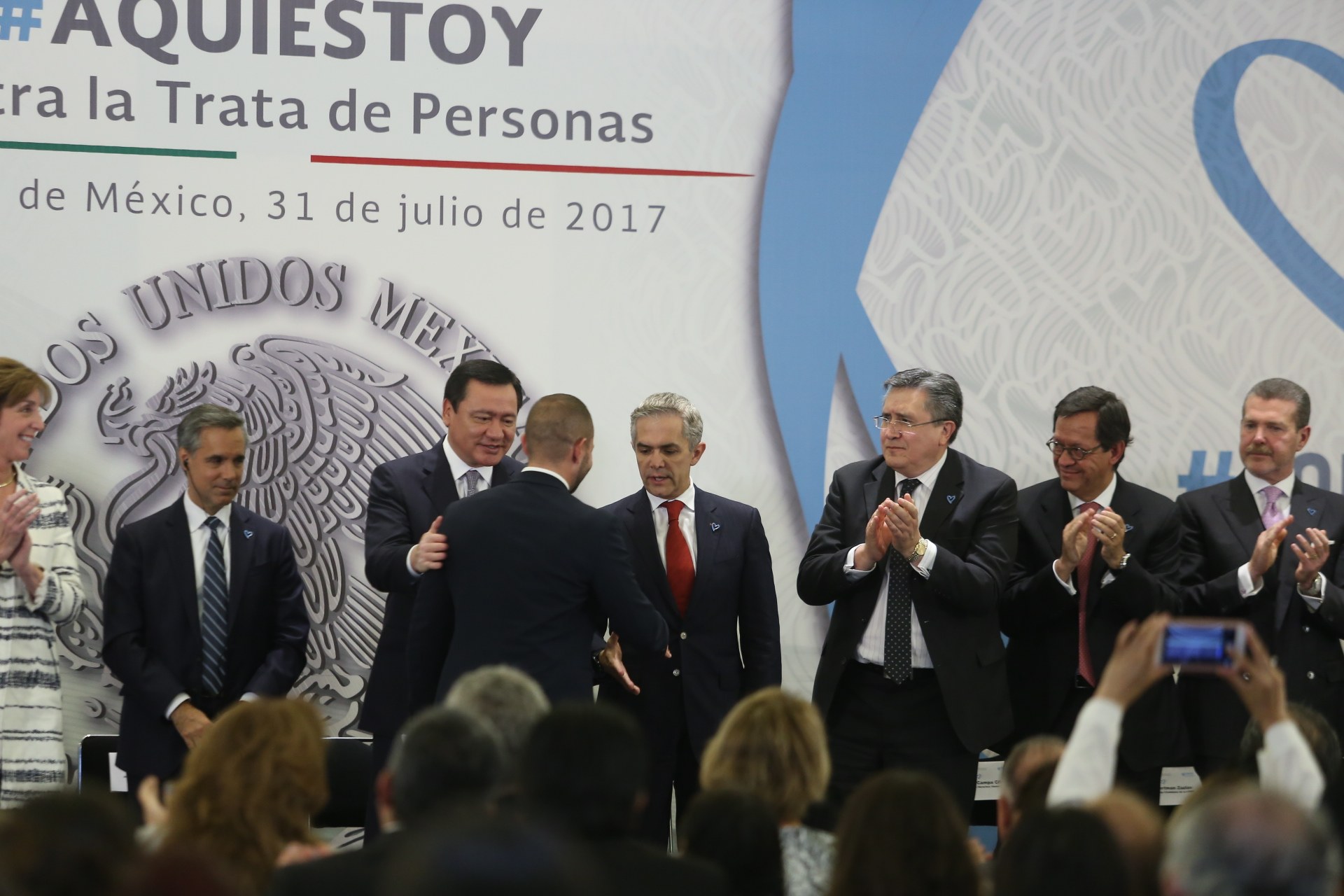 Eine Gruppe von Menschen in Anzügen und legerer Kleidung steht vor einer Menge, einige klatschen, mit einer Person, die ein Handy in der Vordergrund hält und einem Banner mit Text und einem Bild im Hintergrund.