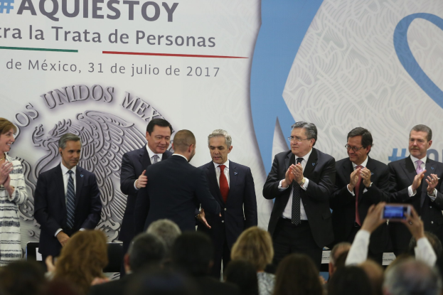 Eine Gruppe von Menschen in Anzügen und legerer Kleidung steht vor einer Menge, einige klatschen, mit einer Person, die ein Handy in der Vordergrund hält und einem Banner mit Text und einem Bild im Hintergrund.