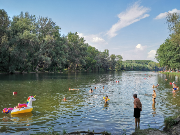 Menschen beim Schwimmen in einem See mit aufblasbaren Spielzeugen, umgeben von grünem Gras, Bäumen, einem hellblauen Himmel und einer Person am Ufer.
