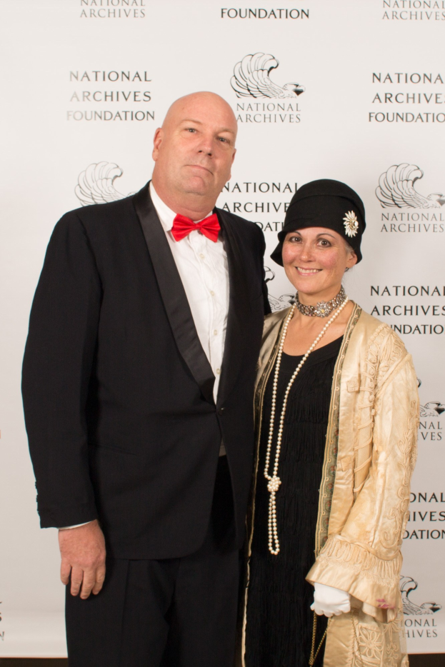 Formalporträt eines Mannes in einem schwarzen Anzug mit weißem Hemd und schwarzer Fliege und einer Frau in einem eleganten Kleid mit einem gemusterten Schal, die nebeneinander auf einem roten Teppich mit einem weißen Banner im Hintergrund stehen.