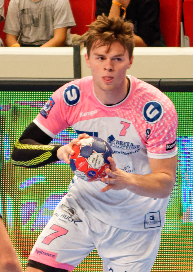 Ein Mann in einem pinken Hemd hält einen Fußball, wobei die Hand einer anderen Person auf der linken Seite zu sehen ist, und im Hintergrund sitzen ein paar Menschen auf Stühlen.