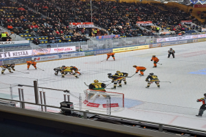 Eine Gruppe von Menschen, die Hockey auf einem Eisplatz mit einem Netz und einem umgebenden Zaun spielen, Zuschauer im Hintergrund und Bannern mit Text, unter Stadionlichtern.