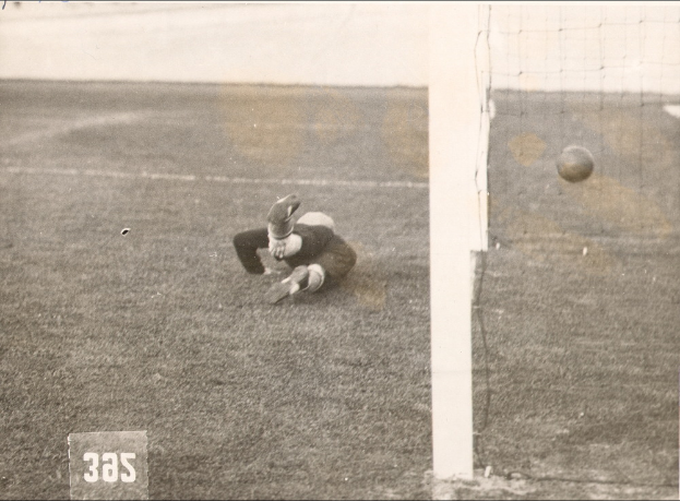 Ein Schwarz-Weiß-Foto eines Fußballspielers, der einen Ball vor einem Tor mit Netz auf der rechten Seite fängt und unten Text.