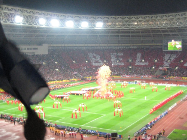 Ein Stadion mit Flutlicht, Eisengittern, Zuschauern, stehenden Personen, Werbetafeln, Bildschirmen, Überwachungskameras und dekorativen Flaggen.