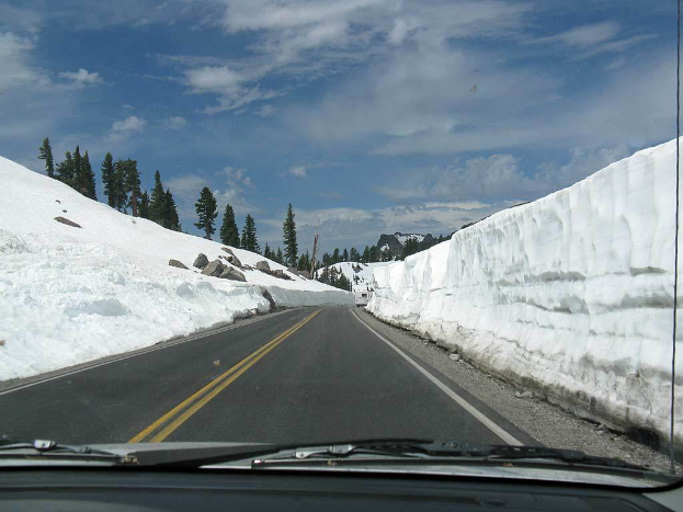 Eine Aufnahme aus einem Fahrzeug, die eine leere Straße mit schneebedeckten Bergen und Bäumen daneben zeigt.