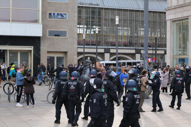 Eine Gruppe von Polizisten in Helmen steht vor einer Menge, die Fahrräder hält, mit Glasfrontgebäuden, Laternenmasten und Fahrzeugen im Hintergrund.