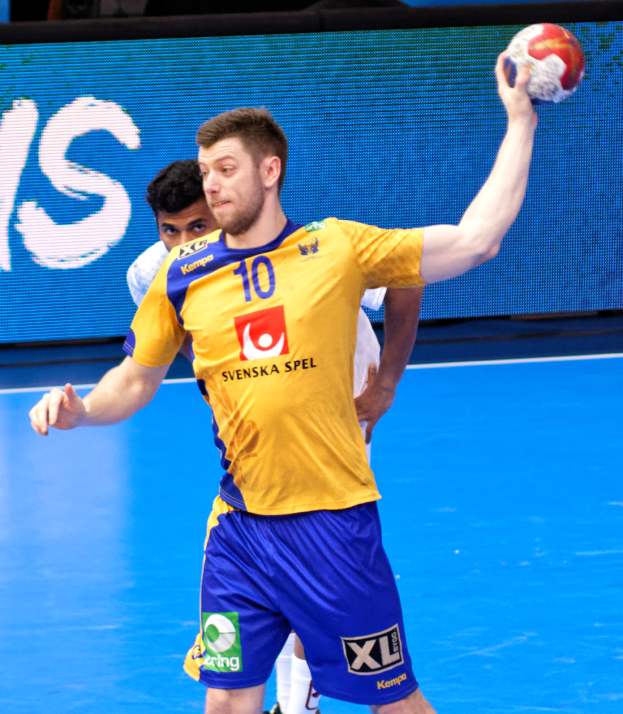 Ein Mann in einem gelben Hemd und blauen Shorts, der auf einem Handballfeld spielt und einen Ball hält, mit einem Hintergrundbild, das "Svenský Spel Handball World Cup 2019" anzeigt.