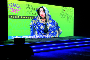 Eine Frau in einem blauen und weißen Kleid steht auf einer Bühne beim World CIO Summit 2022, beleuchtet von hellen Scheinwerfern, mit einem dunklen Hintergrund und einem großen Bildschirm dahinter.