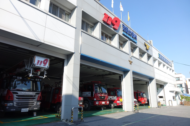 Feuerwache mit mehreren geparkten Feuerwehrautos, einem Gebäude mit Fenstern, Säulen und Namensschildern sowie Flaggen im Vordergrund, vor dem Hintergrund anderer Gebäude und eines klaren blauen Himmels.