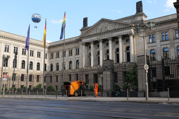 Großes Gebäude mit vielen Fenstern, als Bundestag in Berlin, Deutschland identifiziert, umgeben von Straßeninfrastruktur, Fahrzeugen, Passanten und Bäumen unter einem bewölktem Himmel.
