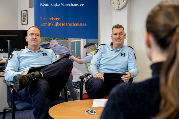 Zwei Polizeibeamte sitzen an einem Tisch mit einer Frau, die Dokumente durchsehen; eine Wanduhr und ein Banner, ein zweiter Tisch mit einem Monitor und Peripheriegeräten sowie Rahmenfotos sind zu sehen.