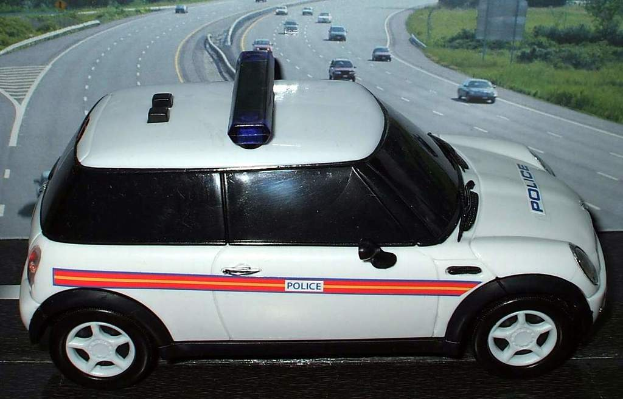 Ein weißer Polizeiwagen ist im Vordergrund geparkt, während mehrere Fahrzeuge auf der Straße dahinter fahren, umgeben von Gras und Bäumen auf beiden Seiten in einer ländlichen Umgebung.