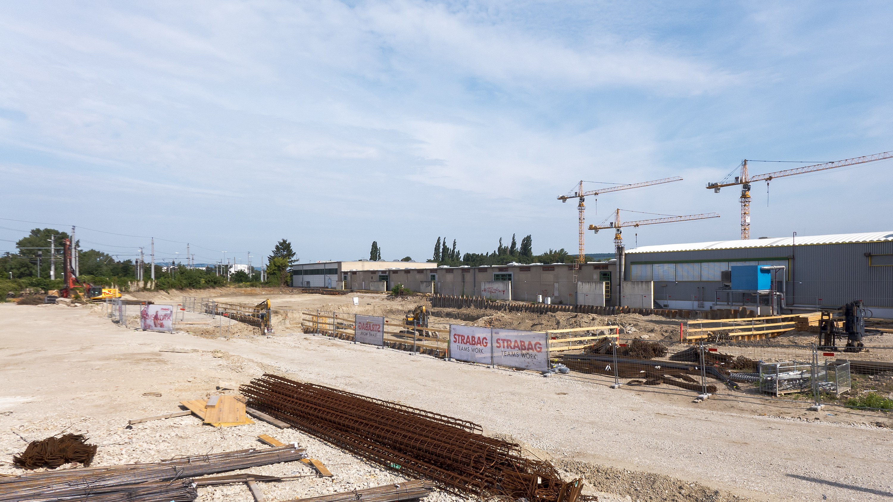 Baustelle mit verschiedenen Materialien und Geräten, darunter Eisenstangen, Holzplanken, Pfähle, Drähte, Kräne und Gebäude, unter einem bewölkten Himmel.