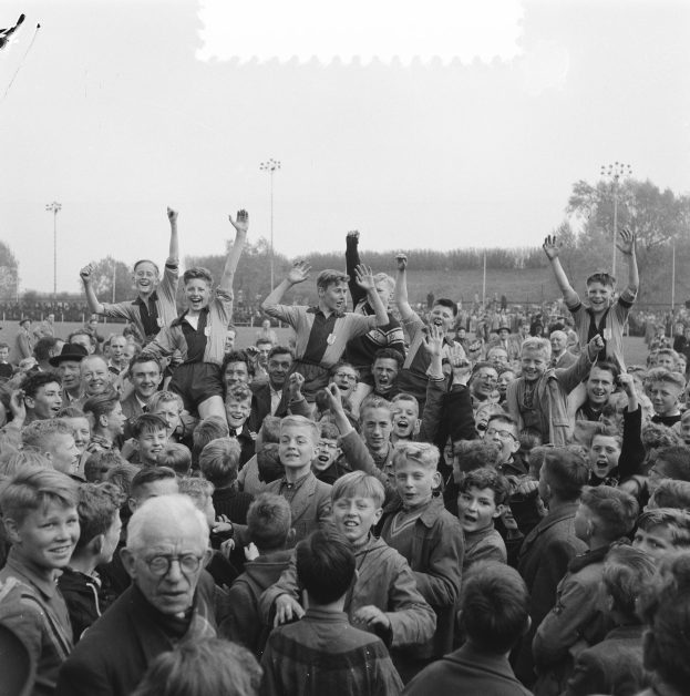 Schwarzes und weißes Foto einer Menge vor einem Stadion, einige feiern mit erhobenen Händen, mit Laternenmasten, Bäumen und einem klaren Himmel im Hintergrund.