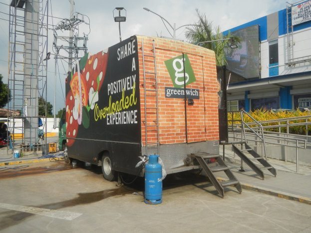 Ein Lkw parkt vor einem Gebäude mit einer Seite, auf der ein Schild mit der Aufschrift "Teilen Sie eine positiv überladene Erfahrung" steht, mit Stufen, die hinaufführen, einem Zylinder auf dem Boden, Geländern, Strommasten mit Drähten, Laternen, Bäumen und einem bewölkten Himmel im Hintergrund.