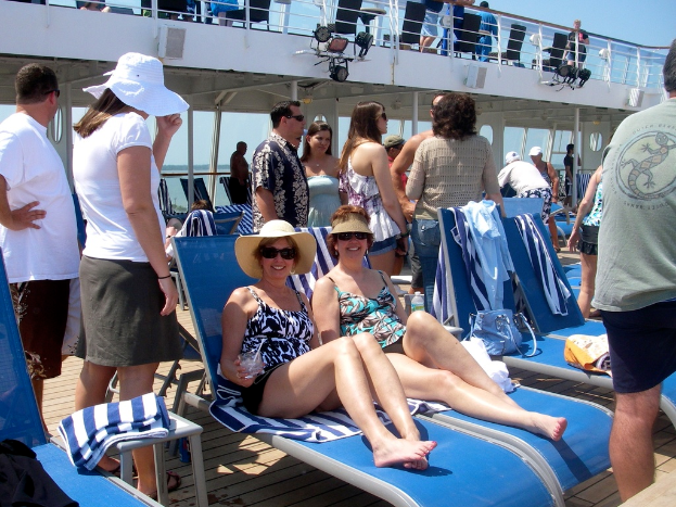 Zwei Frauen in Bikinis sitzen auf Stühlen mit vielen Menschen drumherum unter einem klaren Himmel.