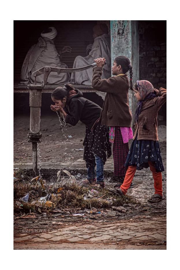 Drei junge Mädchen stehen in der Nähe einer Wasserpumpe in Kathmandu, Nepal; eine trinkt aus dem Hahn, während die anderen warten, mit zwei Personen im Hintergrund auf einer Bank sitzend.