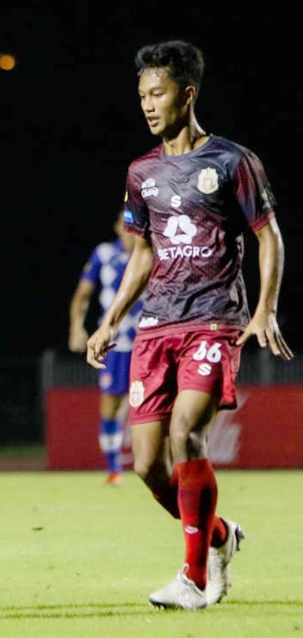 Ein professioneller Fußballspieler in einem marinefarbenen T-Shirt und roten Shorts, der einen Ball auf einem Feld kickt, mit einer anderen Person im Hintergrund und einer Tafel mit Text.