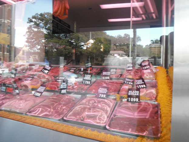 Eine Glasvitrine in einem Fleischgeschäft mit Tabletts voller Fleisch und Preisschildern, mit Gebäuden und Bäumen im Hintergrund und der Decke des Raums oben.