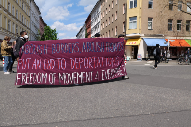 Eine Gruppe von Menschen marschiert auf einer Stadtstraße entlang und hält ein Transparent mit der Aufschrift "Abolish Borders, Abolish Frontiers, Put an End to Deportations, Freedom of Movement 4 Everyone", mit Gebäuden, Bäumen, Fahrrädern und bewölktem Himmel im Hintergrund.
