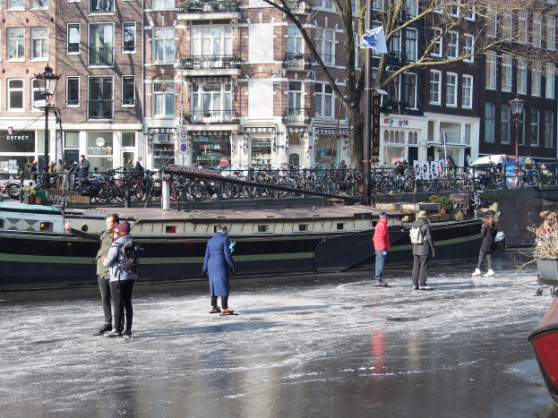Eine Gruppe von Menschen, die auf einem zugefrorenen Kanal in Amsterdam Eislaufen, umgeben von Gebäuden, Bäumen, Laternenmasten, Fahrrädern, Schildern und einer Flagge, mit einem Boot auf der rechten Seite und dem Himmel im Hintergrund.