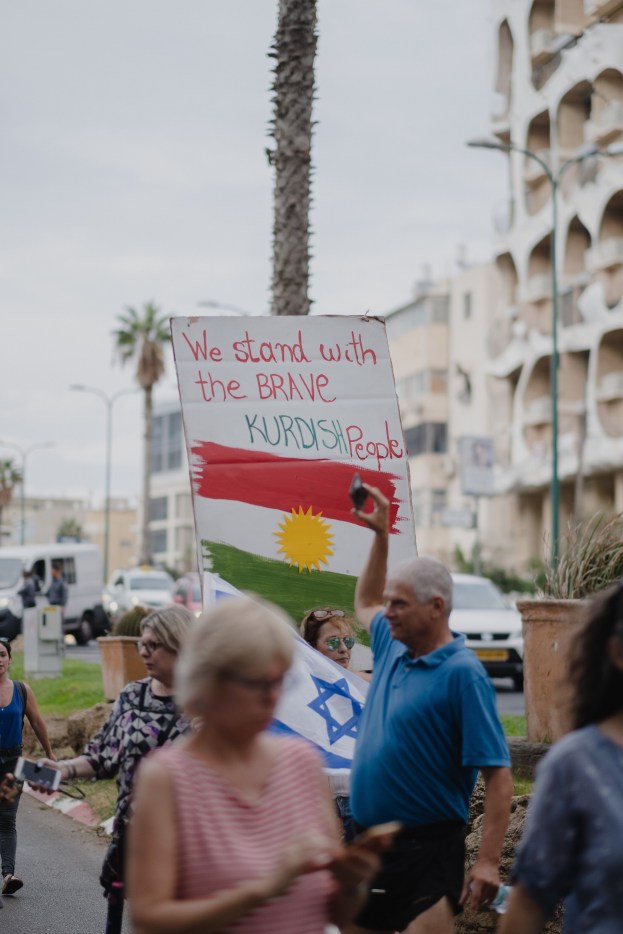 Eine Gruppe von Menschen marschiert nachts eine Straße entlang und hält ein Schild hoch, auf dem steht "Wir stehen zu den mutigen kurdischen Menschen", mit einem Wagen, der am Straßenrand geparkt ist, Straßenlaternen, Bäumen, Gebäuden und einem klaren blauen Himmel im Hintergrund.