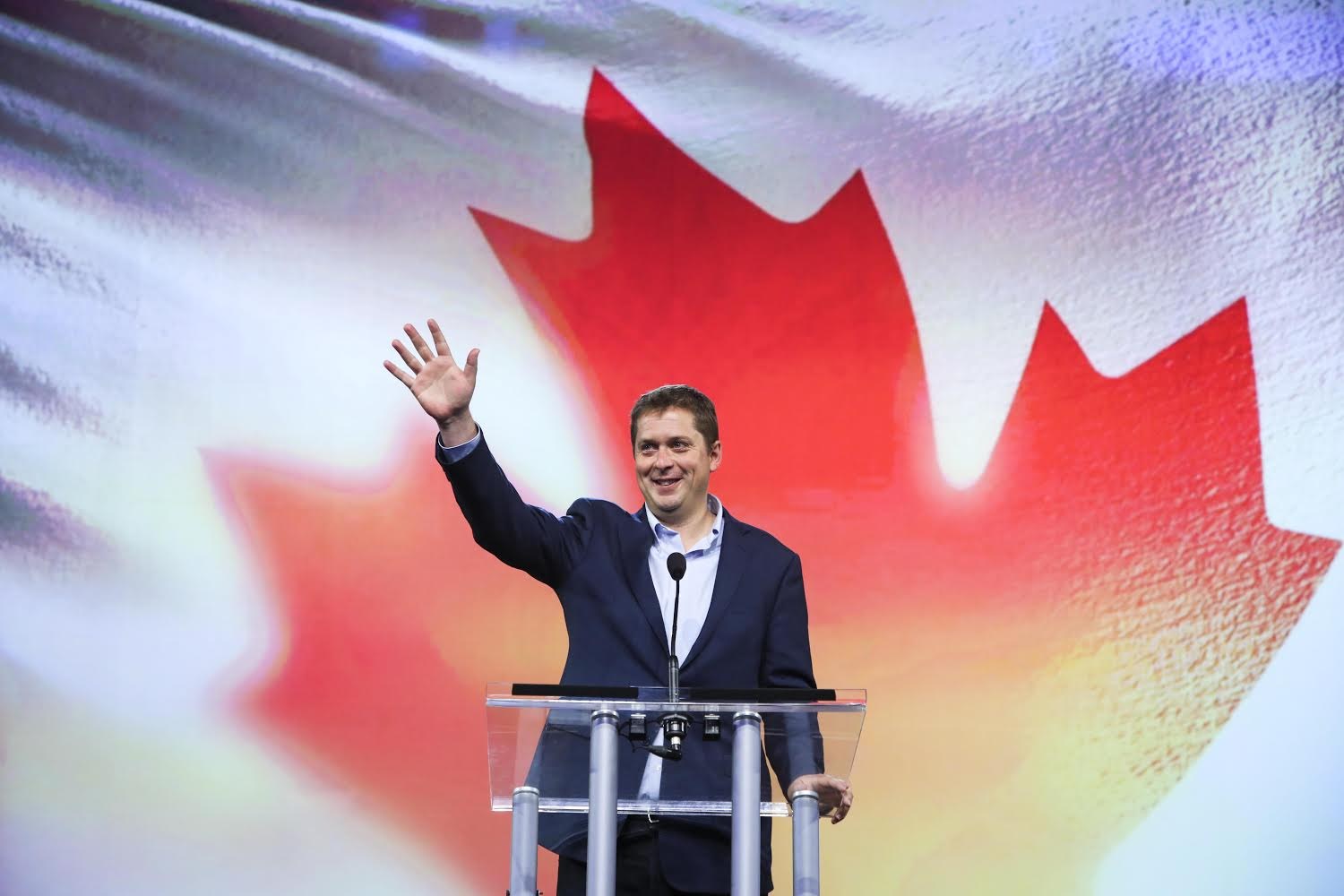 Der kanadische Premierminister Alex Salmond steht an einem Rednerpult mit Mikrofonen, lächelt, vor einer kanadischen Flagge mit einem Bildschirm im Hintergrund.