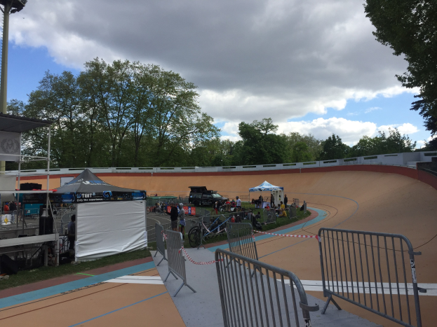 Gruppe von Radfahrern, die auf einer Schmutz-BMX-Strecke bei den UCI BMX-Weltmeisterschaften 2018 fahren, mit Zelten, Fahrzeugen, Geländern, Bäumen und einem bewölkten Himmel im Hintergrund.