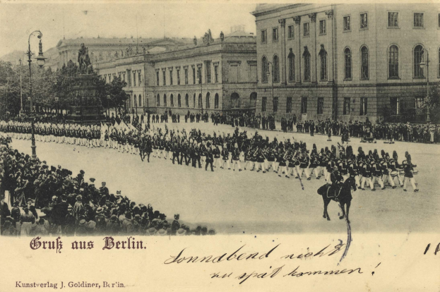 Ein altes Schwarz-Weiß-Foto eines Umzugs in Berlin, Deutschland, das Menschen auf dem Boden und auf Pferden, eine Statue auf einem Sockel, Straßenlaternen, Bäume, Gebäude mit Fenstern und einen bewölkten Himmel zeigt, mit Text am unteren Bildrand.