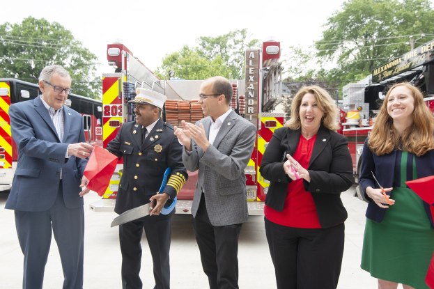 Gruppe von Menschen, die bei einer Einweihungszeremonie der Alexandria Fire Department mit einer Schere und einem roten Band vor einem Feuerwehrauto klatschen und löcheln.