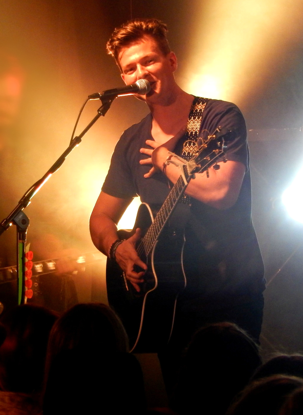 Ein Mann in schwarzem T-Shirt und Jeans spielt Gitarre und singt in ein Mikrofon, während er die Augen schließt und das Publikum begeistert.